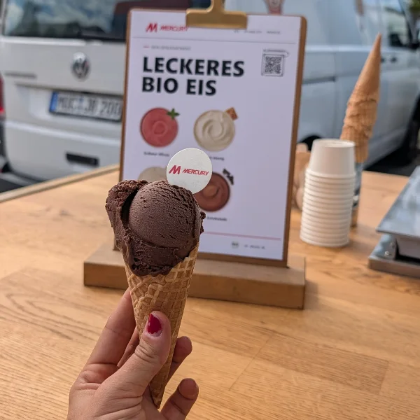 Eine Hand hält eine Waffel mit Schokoladeneis, dahinter die Eiskarte