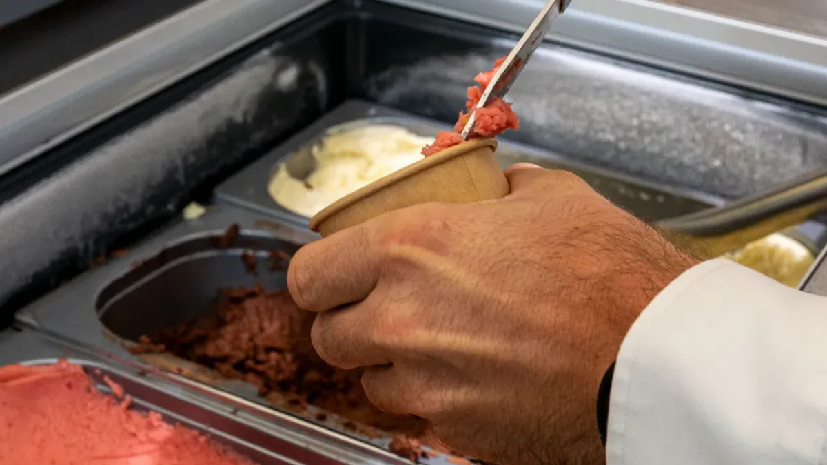Eis wird mit dem Spatel aus der Vitrine in einen Pappbecher gegeben, vorne Erdbeer-Minze, dahinter Schokolade